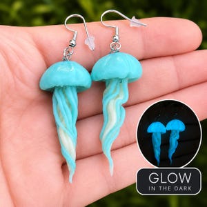 May include: A pair of dangle earrings shaped like jellyfish with light blue translucent caps and swirling white and blue tentacles. The earrings feature silver-toned metal hooks. An inset shows the jellyfish glowing in the dark with the text GLOW IN THE DARK.