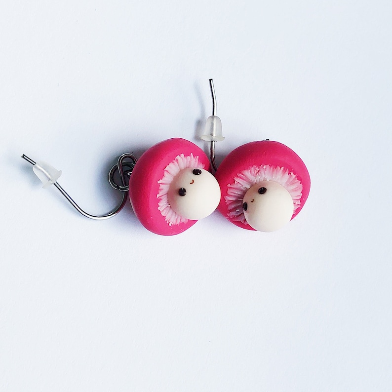 May include: A pair of pink mushroom earrings with white faces and black eyes. The earrings are made of polymer clay and have silver hooks.