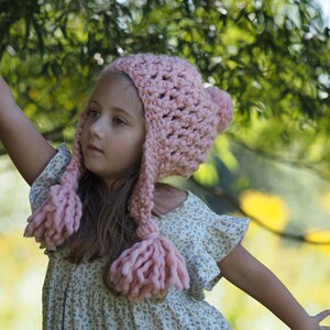 May include: A chunky knit light pink hat featuring a textured open-stitch pattern, a pom-pom on the crown, and two long braided earflaps ending in thick yarn tassels. The hat is crafted from thick, soft yarn and provides full head coverage.