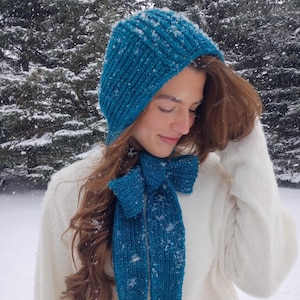Puede incluir: Un conjunto de gorro y bufanda de punto color turquesa. El gorro es de estilo ajustado y la bufanda tiene un detalle de lazo. El conjunto se lleva con un suéter blanco en un entorno nevado al aire libre. El gorro y la bufanda son de hilo suave y texturizado.
