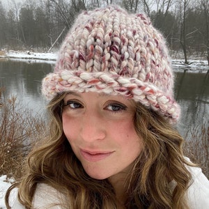 May include: A close-up of a woman wearing a pink and white knitted hat with a wide brim. The hat has a chunky knit texture and is made with a blend of colours.
