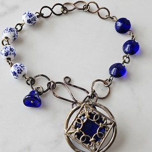 May include: A bracelet featuring a mix of white ceramic beads with blue floral patterns and translucent cobalt blue glass beads. The piece includes a central circular pendant with a filigree frame holding a blue stone and a small blue bell charm.