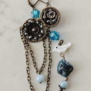 May include: A unique earring with a bronze-colored metal chain and accents. The earring features a circular filigree design, blue beads, a white bird charm, and a dark green stone. The earring has a lever back closure.