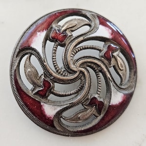 May include: A round, antique button with a silver-colored metal filigree design. The button features a swirling pattern with red and white enamel accents. The outer edge is a deep red color.