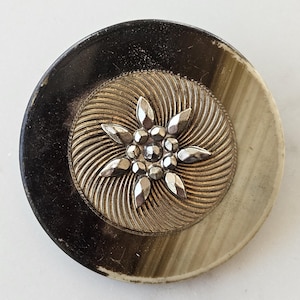 Puede incluir: Un botón redondo, negro y blanco con un diseño de estrella plateada en el centro. La estrella tiene un centro elevado y texturizado y está rodeado por un patrón de espiral.