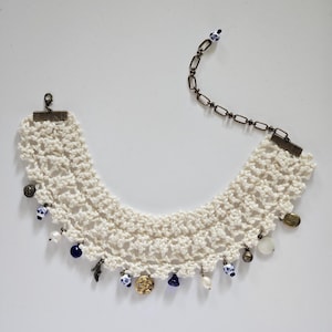 May include: Cream-colored crochet necklace with a variety of charms. The necklace features small blue and white ceramic beads, gold-toned coins, and small pearl-like beads. A dark metal chain extends from the necklace.