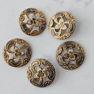 Puede incluir: Cinco botones antiguos de latón con diseño floral. Cada botón presenta un borde dorado de estilo filigrana que rodea una flor central y acentos curvos en metal más oscuro y deslustrado. Los botones son redondos y tienen una estética vintage.