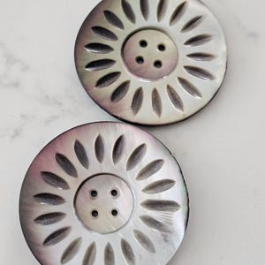 May include: Two large, round buttons with a mother-of-pearl finish. Each button has a central area with four holes and a sunburst design around the edge. The buttons have a pearlescent sheen with hints of grey and purple.