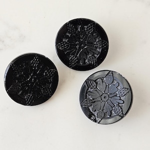 May include: Three black, round buttons with a floral design. Each button features a raised, detailed pattern of petals and leaves radiating from a central point. The buttons are made of a shiny, dark material.