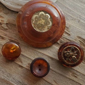 Puede incluir: Cuatro botones vintage de color marrón y dorado. El botón más grande tiene un diseño dorado en relieve en el centro. Los otros tres botones tienen un diseño dorado más sutil.