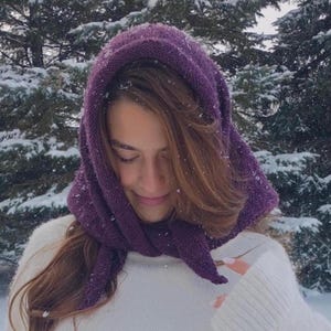 May include: A knitted purple hood scarf, worn over a white sweater, with a snowy forest background. The scarf is a deep shade of purple and appears to be made of a soft, warm material. The image is taken outdoors in a winter setting.