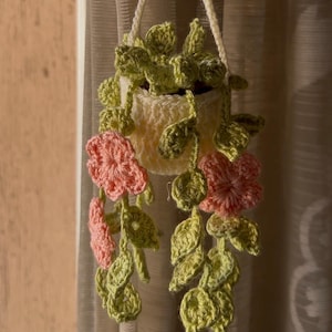 Puede incluir: Un macetero colgante de ganchillo hecho a mano, de color crema, con una cuerda para colgar a juego. El macetero está adornado con hojas verdes y flores rosas. Diseñado para contener pequeñas plantas, añade un toque decorativo.
