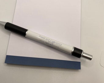 May include: A white notepad with a blue line at the bottom. The notepad has the text "Travel and Experience" printed on the top and the text "Discover, Explore and Experience" printed below it. A black and silver pen with the same text printed on it is lying on top of the notepad.