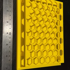 Op de afbeelding: Een geel plastic paneel in de vorm van een honingraat met een verhoogde rand. Het paneel is ongeveer 20 cm breed en 15 cm hoog.