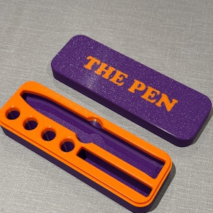 Puede incluir: Un portaplumas morado y naranja con las palabras "THE PEN" impresas en la tapa. El portaplumas tiene seis ranuras para bolígrafos y una ranura para un bolígrafo más grande.