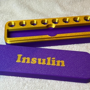Op de afbeelding: Een paarse en gouden insuline-etui met een deksel. Op het deksel staat het woord "Insuline" in gouden letters. De etui heeft een compartiment met zes ronde openingen. De etui is gemaakt van een textuurrijk materiaal.