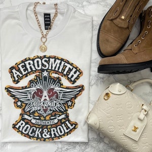 May include: White t-shirt featuring a graphic print of the Aerosmith logo in a leopard print design. The logo includes the text "Aerosmith", "Authentic", and "Rock & Roll".
