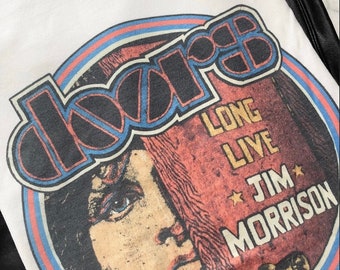 May include: White t-shirt with a vintage-style graphic of the band "The Doors" in a blue and red circle. The graphic features a portrait of Jim Morrison with the text "Long Live Jim Morrison".