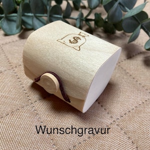 Könnte beinhalten: Eine hellfarbene Holzbox mit gewölbter Ober- und Unterseite, die eine Geldsack-Grafik mit Dollarzeichen zeigt. Die Box ist mit einer braunen Kordel und einem Holzknopf verschlossen. Der Text "Wunschgravur" ist aufgedruckt.