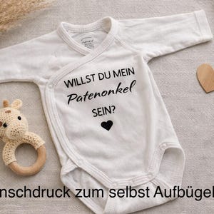 Könnte beinhalten: Weißer Baby-Body mit langen Ärmeln, mit dem Text "WILLST DU MEIN Patenonkel SEIN?" in Schwarz und einem schwarzen Herz. Ein gehäkeltes Giraffenrassel und ein Holzherz sind ebenfalls vorhanden.