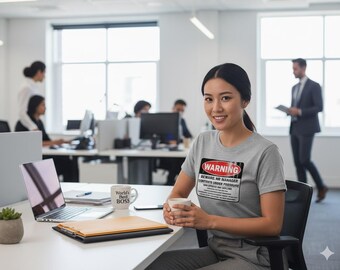 May include: A woman in a gray t-shirt with a warning label that reads "Beware HR Manager Contents Under Pressure". She is seated at a desk with a laptop, mug, and documents. The office setting includes other people and computers.