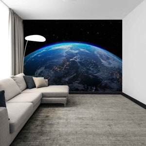May include: A large wall mural depicting a vibrant image of Earth from space, showcasing the blue planet with visible continents and city lights. The mural is set against a black starry background, creating a dramatic and immersive effect. The room features a gray sectional sofa and a gray rug.