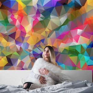 May include: A woman sits on a bed with a white headboard and a colourful geometric patterned wall behind her. She is holding a white fluffy pillow in front of her.