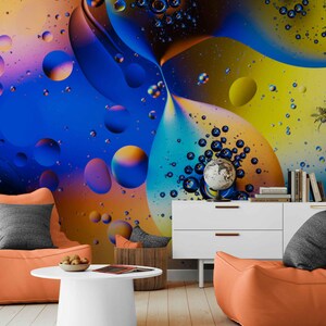 Peut inclure: Un salon moderne avec des poufs oranges, une table basse blanche et un meuble blanc. Le mur présente un design abstrait coloré avec des teintes bleues, jaunes et oranges. Un globe et des livres sont sur le meuble.