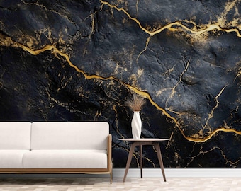 May include: A white sofa sits in front of a wall with a black and gold marble pattern. A small wooden table with three legs stands next to the sofa, holding a white vase containing dried grass. The wall features deep textures and gold veins.