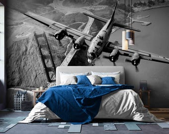 May include: A bedroom featuring a large black and white wall mural of a vintage four-engine bomber aircraft flying over the Golden Gate Bridge. The bed is dressed in white linens with a vibrant blue throw blanket and matching blue decorative pillows.