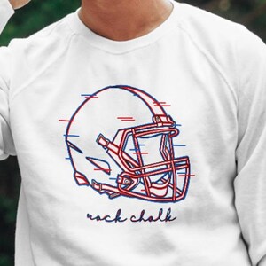 May include: White long-sleeve sweatshirt with a red and blue graphic of a football helmet and the text "rock chalk".