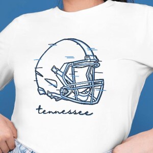 May include: White long-sleeve shirt with a blue football helmet graphic and the word "tennessee" in a script font. The shirt is paired with light-wash denim jeans. The background is a solid blue.