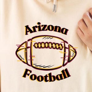 May include: A cream-colored hoodie with a front pocket and drawstring hood. The front features a football graphic with the words "Arizona Football" in black and gold lettering. The football graphic is outlined in black and gold.