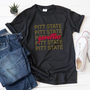 May include: Black t-shirt with the text "PITT STATE" in yellow, repeated vertically, with "gorillas" in red. The shirt is paired with blue jeans and white sneakers, suggesting casual wear.