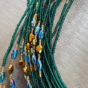 K&ouml;nnte beinhalten: Eine Taillenkette aus gr&uuml;nen und goldenen Perlen. Die Perlen sind auf einer d&uuml;nnen Schnur aufgef&auml;delt und in einem Muster angeordnet. Die Kette ist lang und flie&szlig;end, und die Perlen sind irisierend.