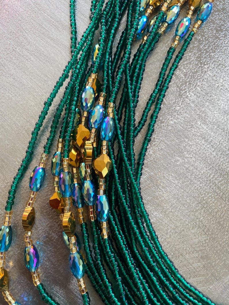 K&ouml;nnte beinhalten: Eine Taillenkette aus gr&uuml;nen und goldenen Perlen. Die Perlen sind auf einer d&uuml;nnen Schnur aufgef&auml;delt und in einem Muster angeordnet. Die Kette ist lang und flie&szlig;end, und die Perlen sind irisierend.