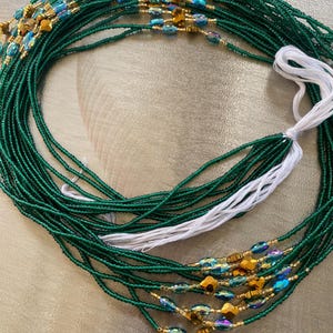 K&ouml;nnte beinhalten: Eine gr&uuml;ne Perlen-Taillenkette mit goldenen und regenbogenfarbenen blauen Perlen. Die Kette besteht aus mehreren Perlenstr&auml;ngen und hat eine wei&szlig;e Schnur daran.