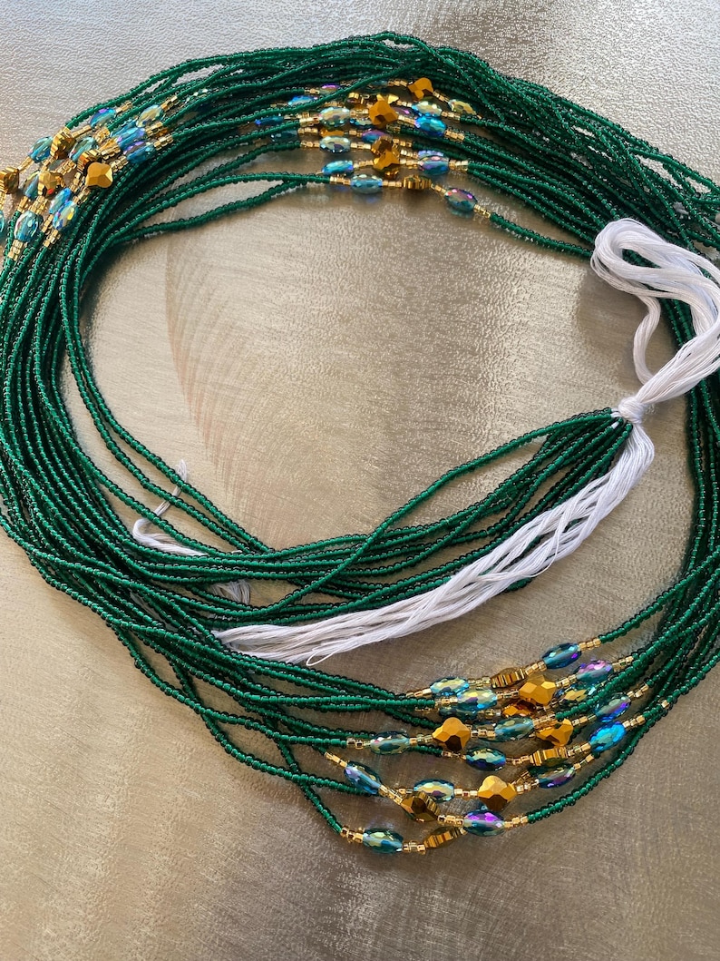 K&ouml;nnte beinhalten: Eine gr&uuml;ne Perlen-Taillenkette mit goldenen und regenbogenfarbenen blauen Perlen. Die Kette besteht aus mehreren Perlenstr&auml;ngen und hat eine wei&szlig;e Schnur daran.