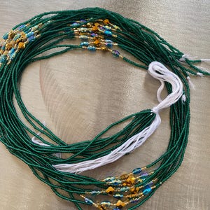 K&ouml;nnte beinhalten: Eine gr&uuml;ne Perlen-Taillenkette mit goldenen und blauen Akzenten. Die Kette besteht aus kleinen Perlen und hat eine wei&szlig;e Schnur daran.