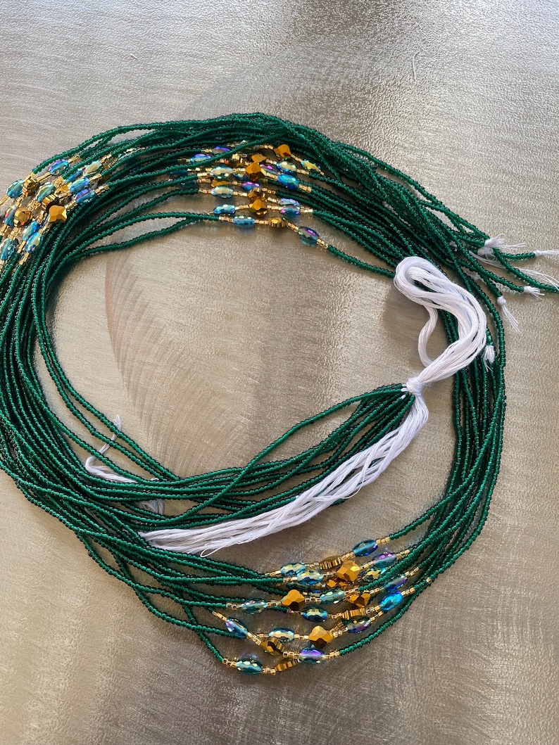 K&ouml;nnte beinhalten: Eine gr&uuml;ne Perlen-Taillenkette mit goldenen und blauen Akzenten. Die Kette besteht aus kleinen Perlen und hat eine wei&szlig;e Schnur daran.