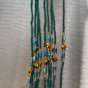 K&ouml;nnte beinhalten: Ein Set aus sieben gr&uuml;nen Perlen f&uuml;r die Taille mit schillernden blauen und goldenen Perlen. Die Perlen sind auf einer d&uuml;nnen Schnur aufgef&auml;delt und etwa 76 cm lang.