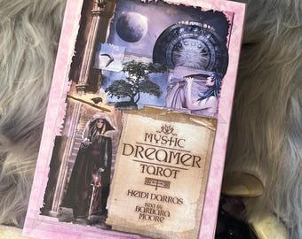 May include: A pink-framed tarot card box titled "Mystic Dreamer Tarot" with celestial and mystical illustrations. Crystals and a white stick surround the box, set on a fluffy grey surface.