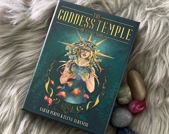 May include: The Goddess Temple Oracle Cards deck box features a green background with gold accents. The box shows a woman with a crown of flowers and a green dress holding a green leaf. The text on the box reads "The Goddess Temple Oracle Cards" and "Sarah Perini & Elena Albanese".
