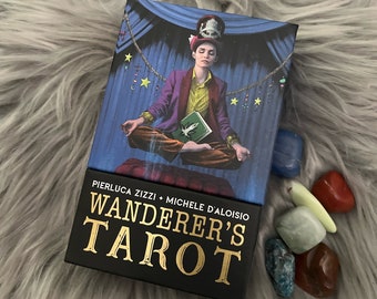 以下が含まれることがあります： Pierluca Zizzi と Michele D'Aloisio による「Wanderer's Tarot」と書かれた金色の文字が書かれた黒い箱。箱には、本を膝に乗せて足を組んで座っている人のカラフルなイラストが描かれています。