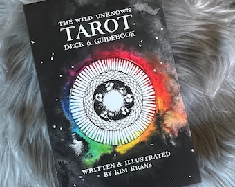 以下が含まれることがあります： The Wild Unknown Tarotのデッキとガイドブック。表紙は黒色で、白い文字とカラフルな円形デザインが施されています。デッキはKim Kransによって書かれ、イラストが描かれています。デッキはふわふわの灰色の表面に置かれています。