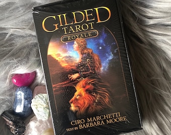 以下が含まれることがあります： 金色の文字と女性とライオンのアートワークが施された黒いGilded Tarot Royaleデッキ。デッキは、ふわふわの灰色の表面にカラフルなクリスタルで囲まれています。