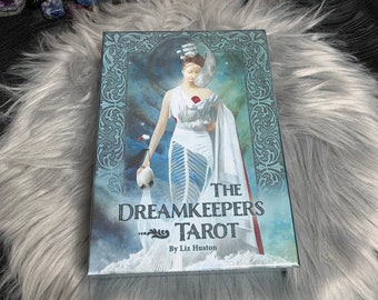 以下が含まれることがあります： Liz HustonのThe Dreamkeepers Tarotデッキ。箱には、赤いバラ、三日月、船を着た白いドレスの女性が描かれています。デッキは、キャンドルとクリスタルが置かれたふわふわの灰色の表面にあります。