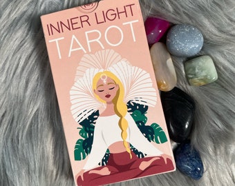 Könnte beinhalten: Eine rosa und weiße Tarot-Deck-Box mit dem Text "INNER LIGHT TAROT" auf der Vorderseite. Die Box zeigt eine Illustration einer Frau, die im Lotussitz meditiert, mit einem weißen Oberteil und einer roten Hose. Die Frau hat blonde Haare und trägt eine weiße Blumenkrone.