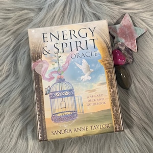 Może przedstawiać: Zapakowana talia kart i przewodnik "Energy & Spirit Oracle" autorstwa Sandry Anne Taylor. Pudełko przedstawia spokojną ilustrację klatki dla ptaków, gołębia i powiewającej tkaniny na tle zachodzącego słońca. Obok talii znajduje się kamień w kształcie gwiazdy i inne kryształy.