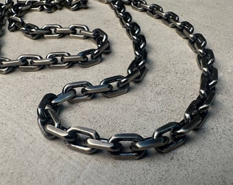 Può includere: Una collana a catena spessa in argento con un design a maglie grandi e quadrate. La catena è appoggiata su una superficie grigia.